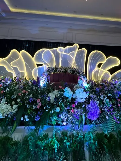 A modern reception stage featuring a large, illuminated, abstract floral structure behind a couch overflowing with flowers.