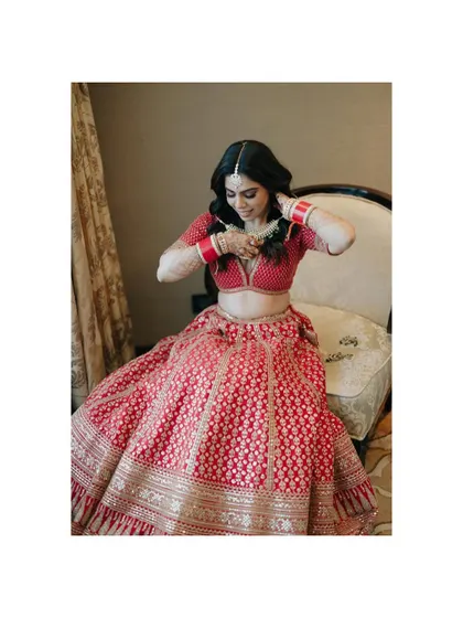 A portrait of the bride getting ready, adjusting her necklace, showcasing the beautiful details of her red lehenga and jewelry.