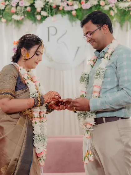 The ring exchange, a pivotal moment in any engagement. The couple is framed by a simple yet elegant backdrop featuring their initials, a soft pink sofa, and a beautiful arch of pastel flowers.