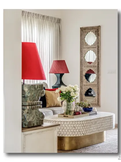 A peek into a living room, where two bold red lamps frame a comfortable sofa. The carved wooden mirror and patterned coffee table add layers of texture and traditional craftsmanship to the space.