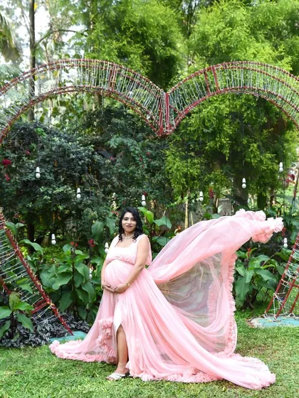 A dramatic outdoor shot featuring a mom-to-be in a flowing pink gown in front of a large heart-shaped arch. The fabric tossing in the wind creates a beautiful, dynamic effect.