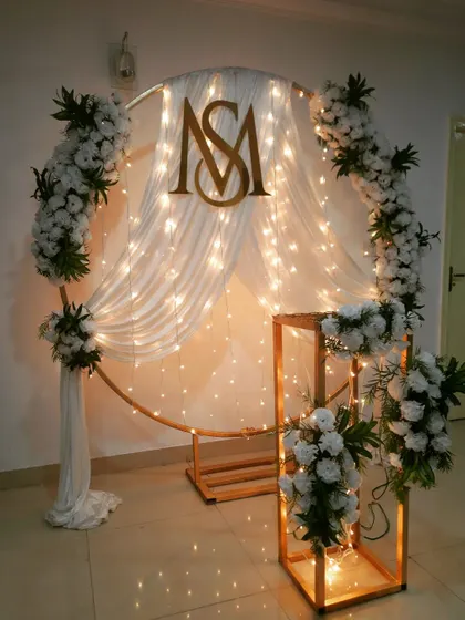 For an intimate wedding anniversary, I designed this modern circular backdrop with white drapes and fairy lights. The custom gold monogram adds a personal touch, and the white floral arrangements complete the elegant look.