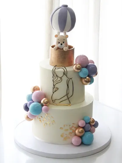 There's nothing sweeter than celebrating a new beginning. This two-tiered baby shower cake features a dreamy hot air balloon with a tiny teddy bear, all handcrafted from fondant, surrounded by a cascade of pastel chocolate spheres. It’s a gentle and elegant way to welcome a little one.