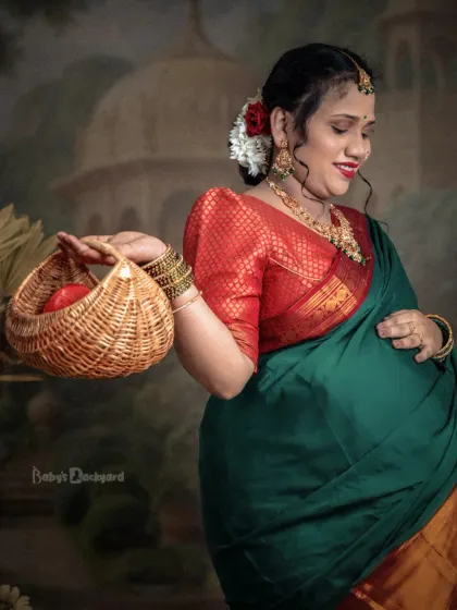 Glowing with happiness in a beautiful green silk saree. The traditional jewelry, flowers in her hair, and a small basket of fruit add to the classic charm of this pregnancy portrait.