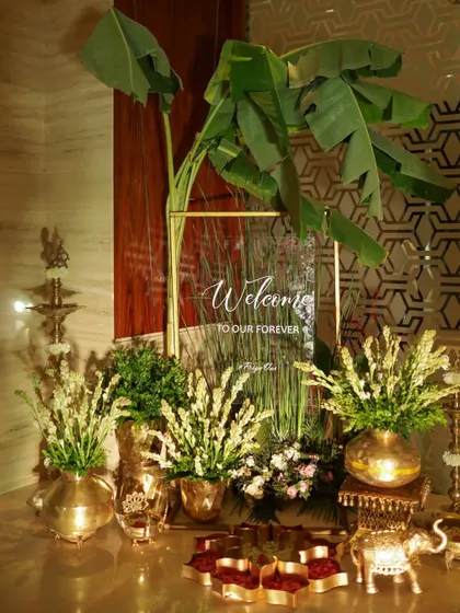 A traditional welcome setup featuring a clear acrylic sign, lush banana leaves, and elegant brass vases filled with tuberoses.