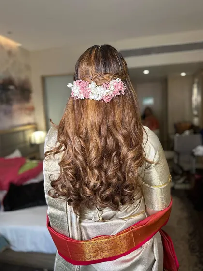 A modern South Indian bridal look with a braided half-updo and soft curls. The hairstyle is adorned with fresh flowers, blending tradition with contemporary style.