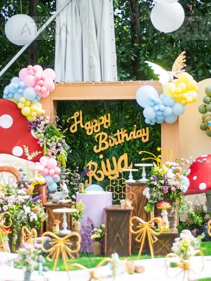 An outdoor woodland-themed birthday party for Riya. The decor includes a lush green backdrop, oversized mushroom props, and whimsical bow-tied chairs, creating an enchanted forest atmosphere for the little guests.