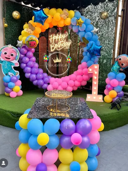 A vibrant Cocomelon first birthday party. This outdoor setup features a rainbow balloon ring, character cutouts, a "Happy Birthday" neon sign, and a light-up number '1'.