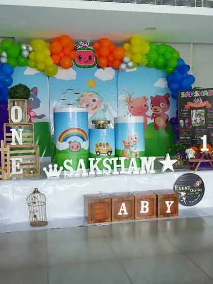 A vibrant Cocomelon setup for Saksham's first birthday. The decor features a rainbow balloon arch and a full cast of character cutouts, creating a playful and colorful scene.