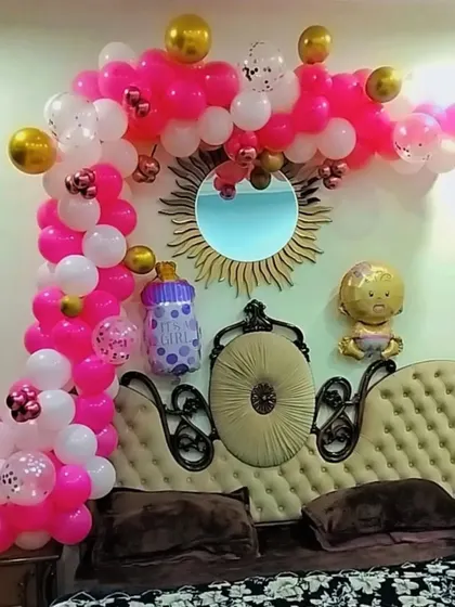 A pink and gold balloon arch over a sofa for a baby girl celebration, with baby-themed foil balloons.