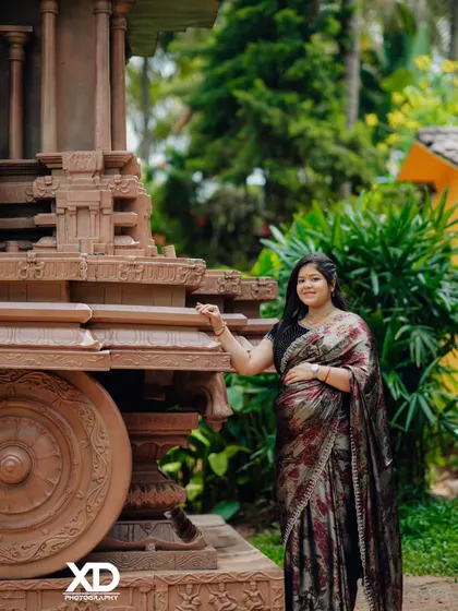 A solo portrait of an expectant mother in a shimmering saree, standing beside a stone chariot, creating a powerful and artistic image.