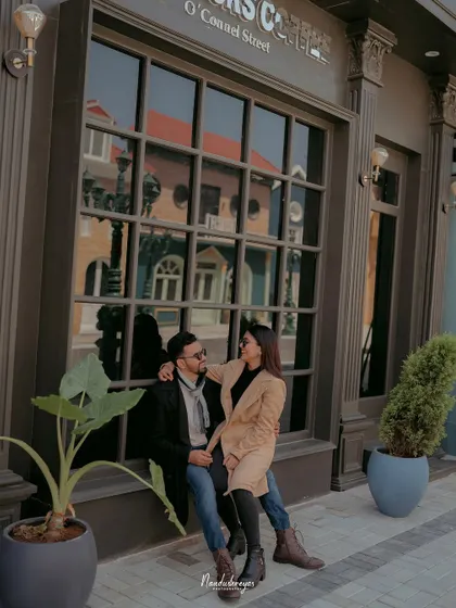 A couple sits together in front of a large window on a city street set, capturing a quiet, urban-cafe-style moment.