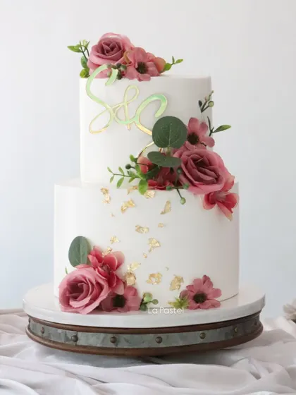 A modern two-tier cake perfect for an engagement or anniversary. The clean white tiers are decorated with deep pink roses, eucalyptus leaves, gold flakes, and a custom initial topper.