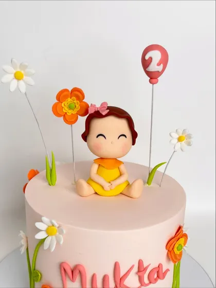 A close-up of an adorable floral-themed cake for a second birthday. The highlight is the sweet, hand-sculpted baby figurine sitting amongst delicate, handcrafted flowers.