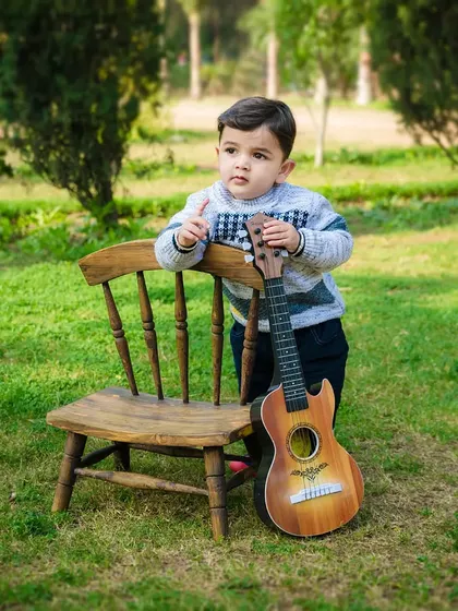 A toddler boy stands next to a small chair and a guitar, looking off into the distance. A cool and casual outdoor portrait.