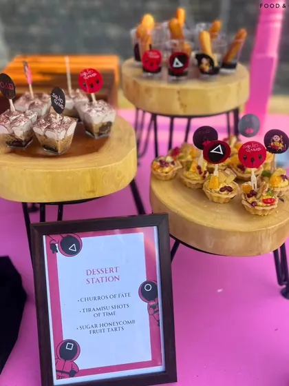 The complete dessert station from our Squid Game party, featuring themed treats like "Sugar Honeycomb Fruit Tarts" displayed on rustic wooden stands.