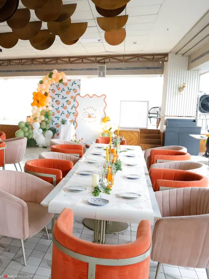 A full view of the baby shower setup, with a long, elegantly set table and comfortable, stylish seating in shades of orange and pink that complement the "cutie" theme.