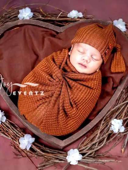 This rustic newborn portrait features a baby swaddled in a warm, russet-colored wrap, nestled inside a heart-shaped wooden bowl. The surrounding twig wreath adds a natural, earthy touch to the composition.