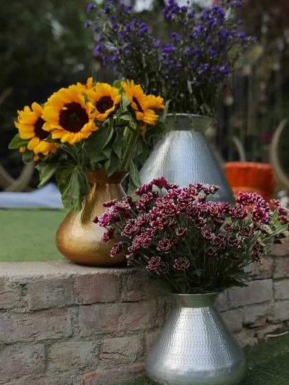 A mix of sunflowers and chrysanthemums in silver and gold vases, placed on a rustic brick ledge.