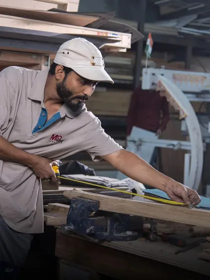 Behind every masterpiece are the hands of our skilled artisans. Here, a craftsman meticulously measures a piece of wood, a critical first step in blending tradition with artistry to create timeless furniture.
