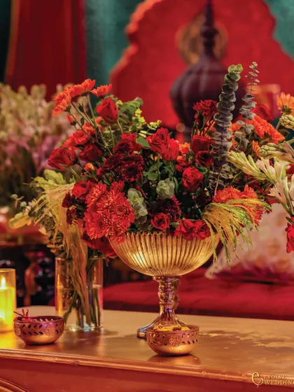A close-up of a floral centerpiece in a golden bowl, with the warm glow of candles creating an intimate atmosphere. The rich red hues of the flowers and seating create a harmonious and luxurious feel.