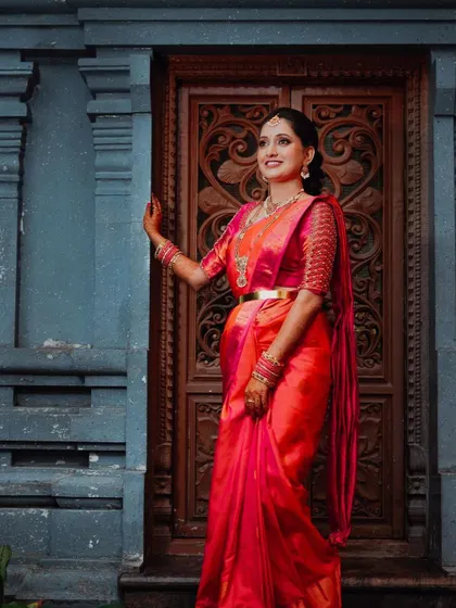 A vibrant portrait of the bride in a stunning red and pink silk saree. The rich colors and the traditional backdrop of a carved wooden door create a culturally beautiful image.