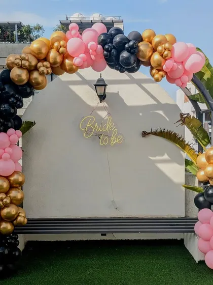 A straight-on view of the 'Bride to be' balloon arch against a white wall on a green terrace, with a neon sign in the center.
