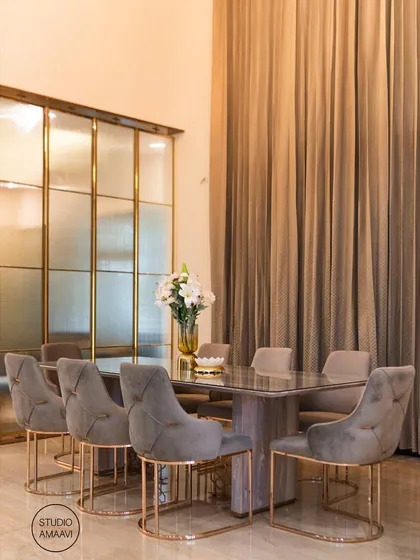 This dining area, part of a luxury villa project, features a double-height ceiling and dramatic floor-to-ceiling curtains. The grey upholstered chairs have elegant rose-gold finished frames, which complement the fluted glass and brass partitions.