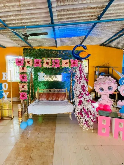A baby shower stage setup in a semi-outdoor space, featuring a decorated swing, a green wall backdrop, and gender-reveal props.