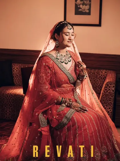 A full-length portrait of the bride, Revati, seated and looking serene in her stunning red lehenga. The soft light beautifully captures the richness of her attire and her calm demeanor.