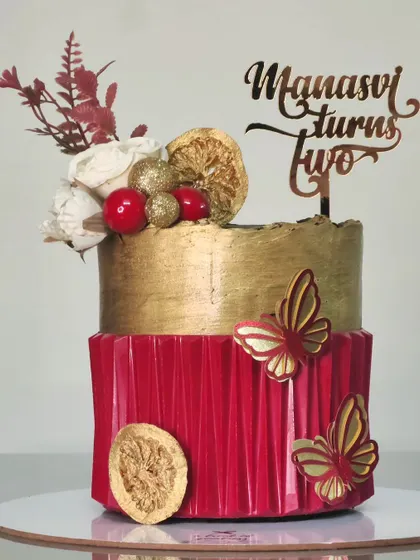 A bold and beautiful two-tiered birthday cake. The bottom tier has a red origami texture, while the top is finished in metallic gold and adorned with dried orange slices and flowers.