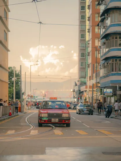 One of Hong Kong's iconic red taxis navigating the tram tracks. The warm, hazy light of the afternoon gives this street scene a timeless, almost vintage feel.