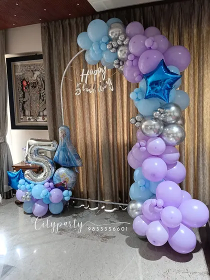 A 'Frozen' themed 5th birthday party setup for a home celebration. The arrangement includes a balloon arch in shades of blue, purple, and silver, with a large '5' foil number and an Elsa character balloon.