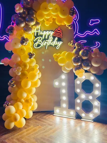 A bright and cheerful 18th birthday decoration. The setup uses a yellow balloon arch, a custom neon sign, and large marquee numbers to mark the special milestone.