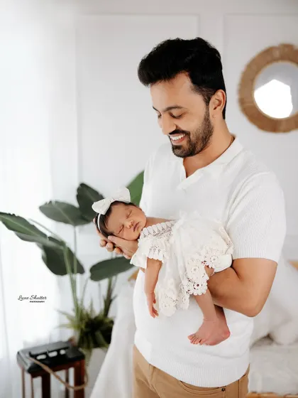 A proud father smiling as he holds his sleeping newborn daughter, who is dressed in a delicate lace outfit.