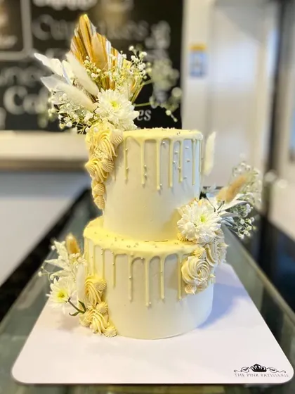 An elegant all-white, two-tiered cake with a delicate white chocolate drip. The cake is adorned with white dried florals, baby's breath, and textured buttercream piping for a sophisticated, monochromatic look.
