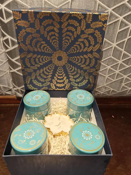 A sophisticated corporate gift set in a deep blue and gold box. Inside are four matching tins for treats like flavored teas or dry fruits, accompanied by a decorative candle.