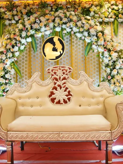 A close-up of a baby shower stage with a dense floral arch of white and pink flowers and an ornate white diwan for the parents-to-be.