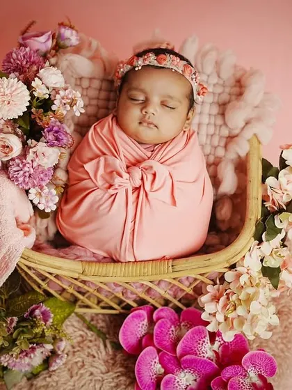 A newborn girl sleeps peacefully in a wicker bassinet, surrounded by a cascade of pink and purple flowers. We use a variety of props to create these enchanting setups.