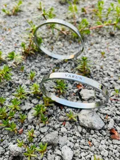 Two silver Kadas, both engraved with the name 'Varun', placed in a natural outdoor setting. This shows how you can order matching bracelets for friends or partners.
