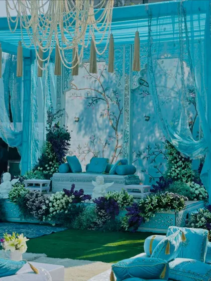 This is my 'Firozi Ishq' theme, a celebration of turquoise. The main seating area features a hand-painted backdrop, framed by flowing turquoise drapes and surrounded by a garden of purple and white flowers.