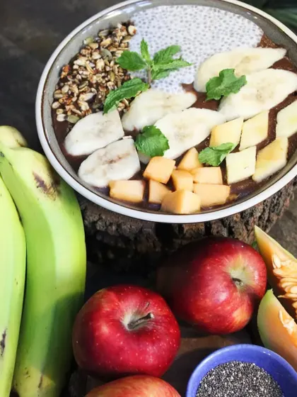 Our smoothie bowl surrounded by the fresh, whole ingredients that go into it: raw bananas, apples, and melon. We believe in using real food to create real nourishment.