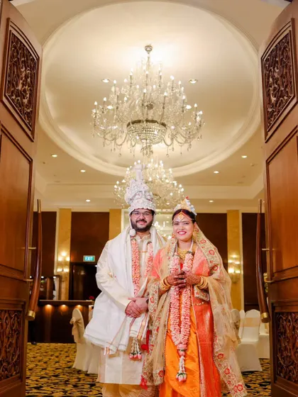 A grand portrait of a Bengali couple in a luxurious hotel lobby. The massive chandelier and ornate doors frame them perfectly, adding to the opulence of the day.
