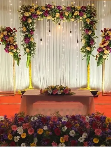 This modern stage design combines a lush floral arch with industrial-style hanging bulbs against a clean white curtain. The setup is framed by matching floral stands, offering a chic and sophisticated look for an engagement or reception.