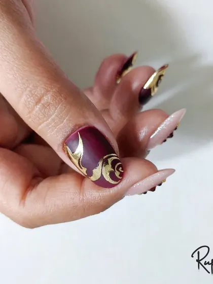 A close-up of the burgundy and gold foil swirl design on a client's hand. The stiletto nail shape adds to the drama.