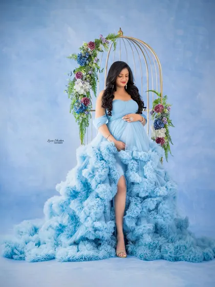 A fairytale moment. The mom-to-be is seated in a golden birdcage adorned with flowers, wearing a magnificent ruffled blue gown.