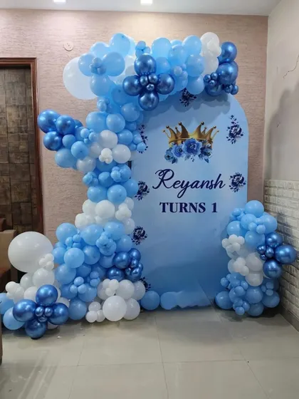 A prince-themed first birthday decoration for Reyansh. The setup includes a blue and white balloon arch over a custom backdrop with a crown motif.