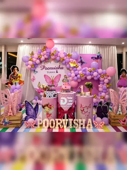 A vibrant butterfly-themed first birthday for Poorvisha. This setup is full of color, with a rainbow-striped floor, purple and pink balloon arches, and numerous butterfly details.