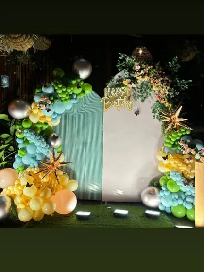 A sophisticated birthday setup in a covered outdoor area. This decoration features dual backdrops and an organic balloon garland in shades of green, yellow, and blue, accented with floral arrangements.