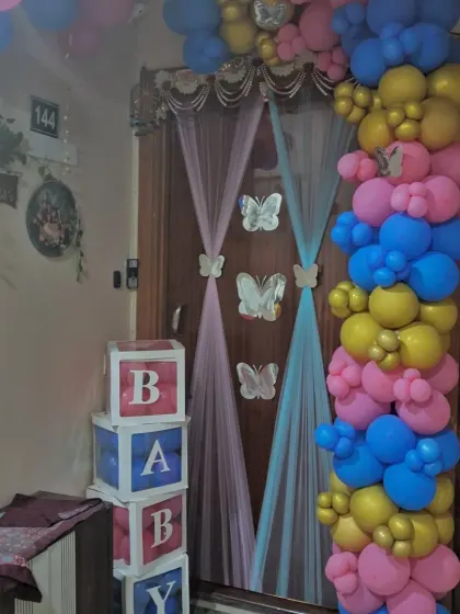 A creative door decoration for a baby shower. This setup uses a colorful balloon garland, sheer curtains with butterfly details, and stacked 'BABY' blocks to welcome the new arrival.